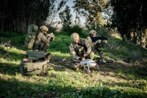 Soldiers in camouflage gear crouch in a field with a white quadcopter-style drone on the ground, packing equipment nearby.